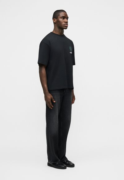 Άνδρας στέκεται φορώντας υπερμεγέθη μαύρο T-shirt με γραφικό στο στήθος, φαρδιά σκουρόχρωμα τζιν και μαύρα αθλητικά παπούτσια σε λευκό φόντο.