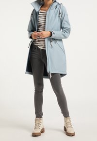 Manteau bleu clair avec capuche, cordons de serrage et fermetures éclair à l'avant. Associé à un t-shirt rayé, un jean gris et des bottes à lacets de couleur crème.