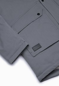 Grå jacka med en slät textur, med knappstängning, sidfickor och en svart etikettdetalj. Standardpassform och modern design.