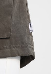 Veste kaki avec une texture légèrement brillante, dotée d'une étiquette latérale, d'un col à revers et d'un ourlet sur mesure. Design simple avec des coutures subtiles.