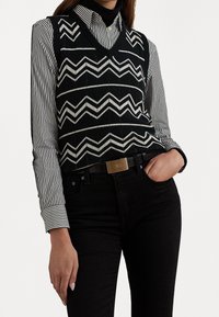 Czarna i biała koszula w paski z długim r ękawem, noszona pod czarną kamizelką z wzorem typu chevron; do tego czarne dżinsy i metaliczny pasek.