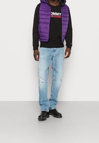 Chaleco morado acolchado sobre un sudadera negra con el logo "TOMMY JEANS", combinado con jeans desgastados de color azul claro y botas negro hasta el tobillo.