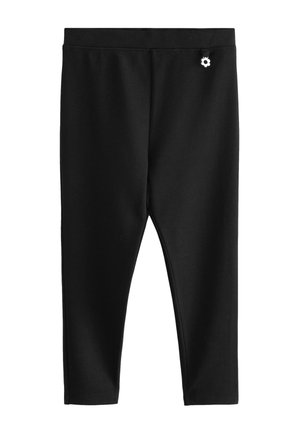 Schwarze eng anliegende Hose mit elastischem Bund und einem kleinen weißen Blumenanhänger nahe der Taille vor einfarbigem Hintergrund.