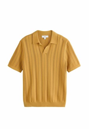 Młody mężczyzna w musztardowożółtych dzianinowych szortach i koszulce polo stoi przy kamiennym murze obok drewnianych drzwi, mając na sobie czarne sandały, okulary przeciwsłoneczne i torbę na ramię.