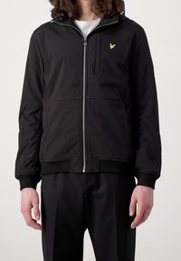 Zwarte bomberjas met capuchon, gemaakt van gladde stof. Voorzien van een ritssluiting aan de voorkant, zijzakken en een klein geel logo op de borst.