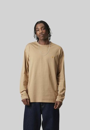 Giovane con capelli intrecciati che indossa una maglia beige a maniche lunghe e jeans blu scuro, in piedi davanti a uno sfondo grigio uniforme.