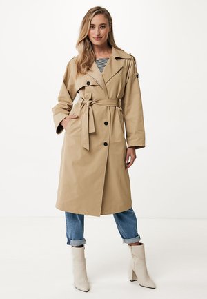 Un abrigo trench beige con cintura ceñida por un cinturón, grandes solapas y botones de doble botonadura. Combinado con unos vaqueros azules arremangados y botas blancas hasta el tobillo.