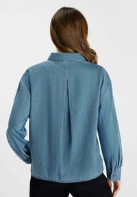 Lichtblauwe denimshirt met lange mouwen, een kraag, een rugplooi en een subtiel gestippeld patroon. Gladde structuur en casual ontwerp.