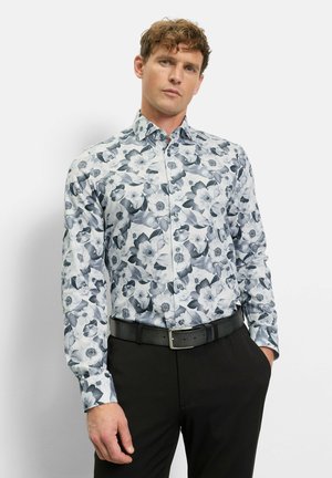 Man draagt een nauwsluitend, lang mouw, knoopjesoverhemd met zwart-witte bloemenprint, zwarte broek en een zwarte riem, staand met één hand in de broekzak.