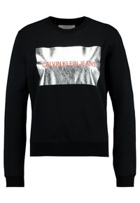 Felpa nera con un pannello riflettente argento con la scritta "CALVIN KLEIN JEANS" in rosso. Polsini e bordo a costine, design dalla vestibilità rilassata.