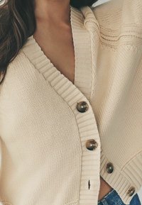 Nahaufnahme einer Person, die eine beigen Strickjacke mit braunen Knöpfen über blauen Jeans trägt.