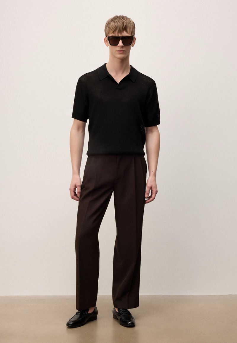 Joven con polo de punto negro de manga corta, pantalones marrón oscuro, mocasines de cuero negro y grandes gafas de sol negras, de pie frente a un fondo liso.