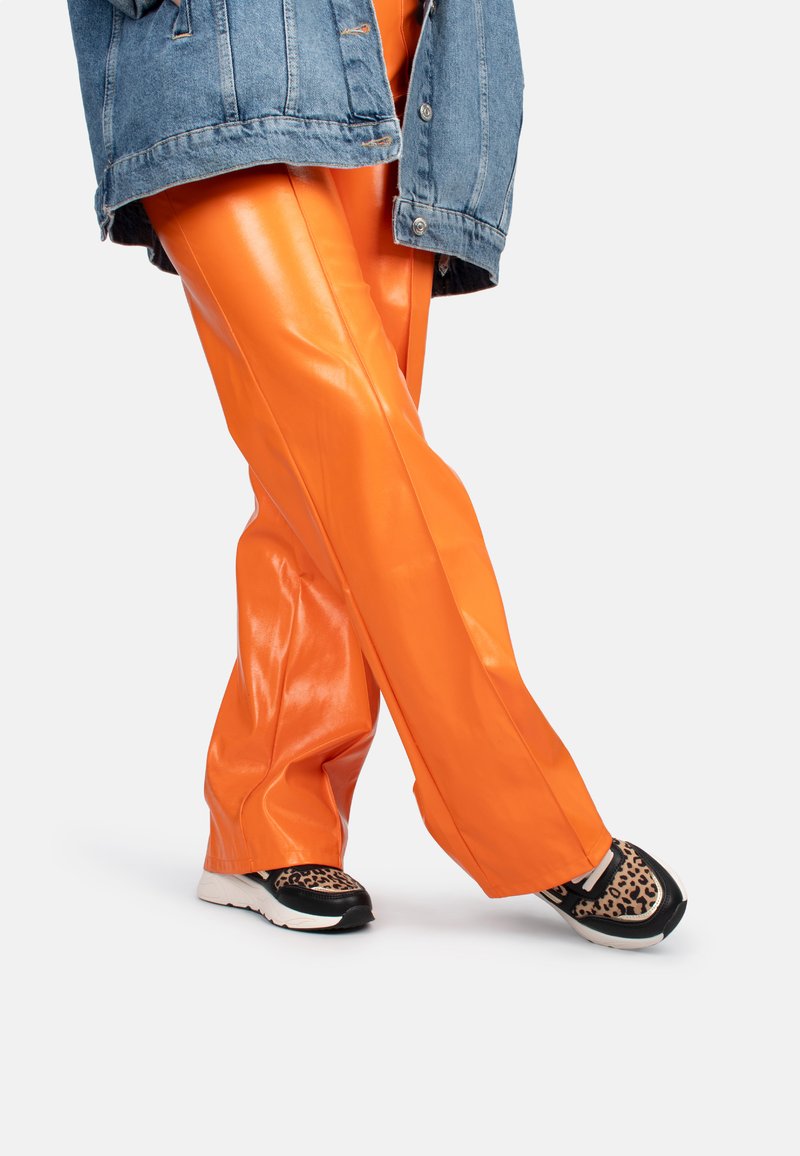 Pantalones de ecológico naranja con un acabado brillante, combinados con zapatillas negras y de estampado de leopardo. Llevando una chaqueta de denim encima.