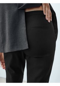 En person i svarta byxor och en grå jacka, med ena handen vilande på höfterna ovanför bakfickan.