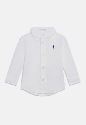 Biała koszulka z długim rękawem dla malucha z kołnierzykiem na guziki i guzikami z przodu, z małym granatowym haftowanym logo gracza polo na piersi.