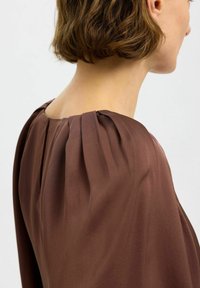 Blusa marrone con maniche a sbuffo, caratterizzata da una texture morbida e dettagli plissettati sulla spalla. Il retro presenta un sottile scollo a barca.