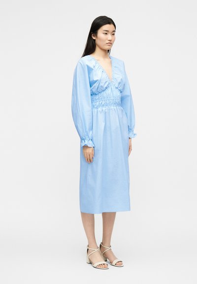 Νεαρή γυναίκα φοράει ένα ανοιχτό μπλε μεσαίο φόρεμα με μακριά φουσκωτά μανίκια και μπεζ πέδιλα με φαρδιά τακούνια και λουράκι στον αστράγαλο σε λευκό φόντο.