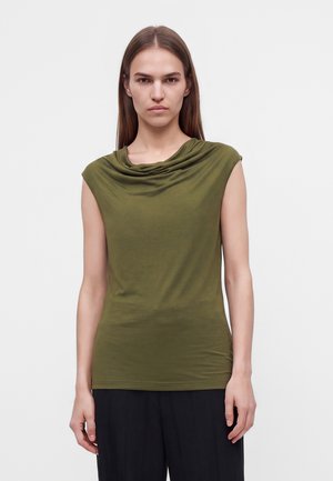 Un mannequin se tient devant un fond uni, vêtu d’un haut sans manches vert olive au décolleté drapé et d’un pantalon noir.