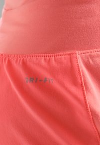 Tela de poliéster coral con textura suave. Presenta un logo DRI-FIT en un gris contrastante, con costuras visibles en el dobladillo.