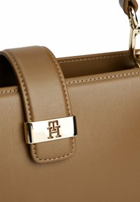 Torebka skórzana w kolorze tan, charakteryzująca się gładką fakturą, usztywnioną konstrukcją i złotym akcentem logo na pasku.