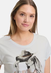 T-shirt en coton bleu clair présentant un graphique d'une femme portant un chapeau à larges bords, orné d'accents métalliques et de perles colorées.