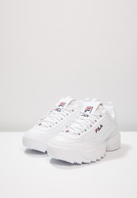 Baskets de sport blanches avec un dessus en cuir texturé, semelle en caoutchouc épaisse, et logo Fila en rouge et bleu sur la languette et sur le côté.