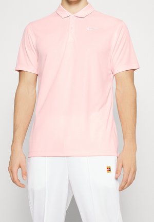 Mann trägt ein hellrosa Nike Polohemd und weiße Hose mit einem kleinen bunten Aufnäher am Oberschenkel, steht vor einem einfarbigen Hintergrund.