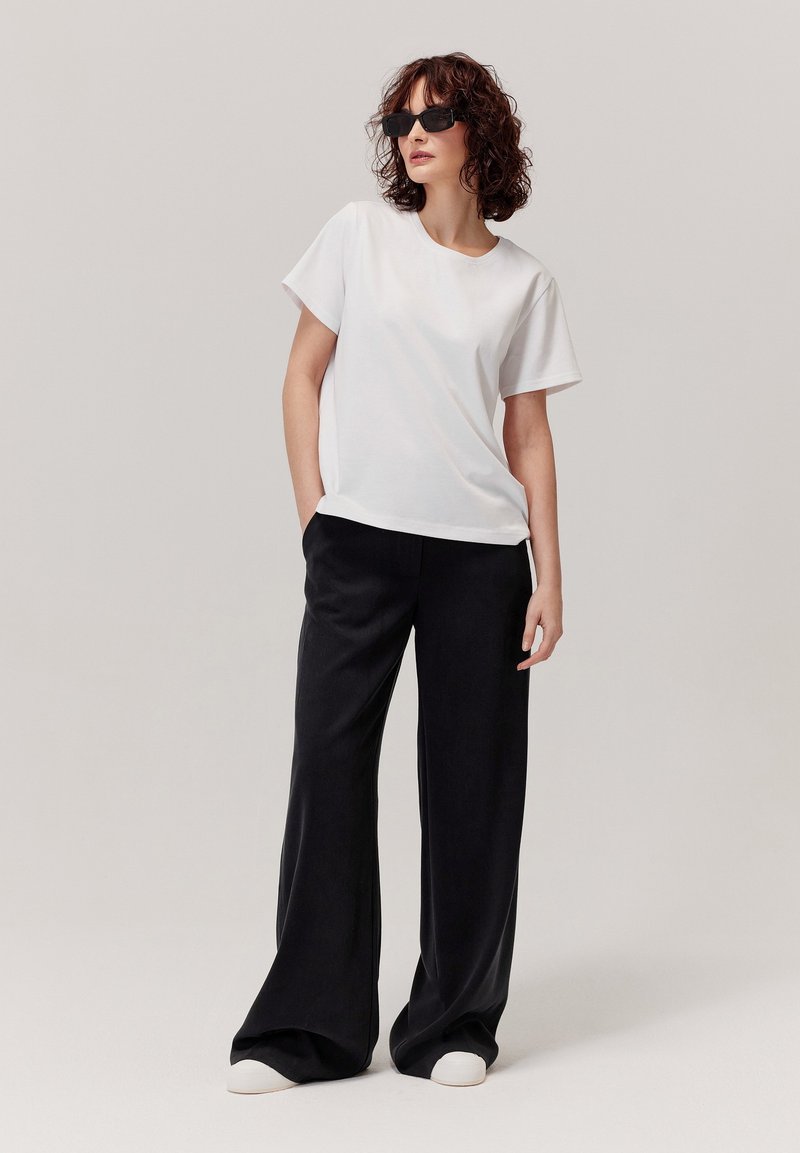 Biała koszula z krótkim rękawem, czarne szerokie spodnie i białe trampki. Modelka w czarnych okularach przeciwsłonecznych, z kręconymi włosami ułożonymi w stylu.