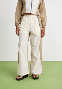 Conjunto deportivo ligero en tonos beige y crema, que incluye una chaqueta corta con cremallera y pantalones de pierna ancha con cinturilla elástica y bolsillos laterales.