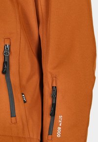 Rostorange Jackenstoff mit zwei vertikalen schwarzen Reißverschlüssen und dem in weißer Stickerei auf dem Ärmel aufgestickten STX 8000-Logo.