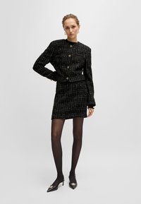 Mustat tweed-puku, jossa on istuvat takki ja mini-hame, viimeistelty kultaisilla napeilla. Mallilla on päällä läpinäkyvät mustat sukkahousut ja teräväkärkiset mustat korot.
