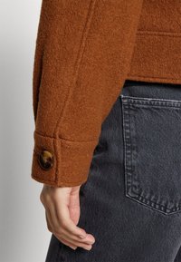 Manga de chaqueta marrón texturizada con puño abotonado usada sobre vaqueros negros de mezclilla, mostrando el bolsillo trasero y la mano descansando al lado.