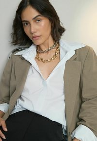 Beiger Blazer über einem weißen Hemd, der mit großen Gold- und Silberketten verziert ist. Dunkle Hosen runden das Outfit ab. Nahaufnahme-Porträt.