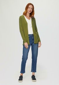 Groene gebreide cardigan met een open voorkant, gecombineerd met een wit V-hals T-shirt en blauwe jeans. Model draagt zwarte sneakers. Soepele textuur.