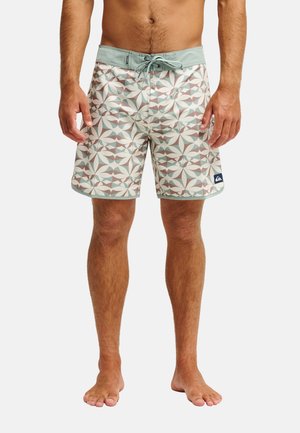 Homme portant un short de bain à motifs avec une ceinture et un cordon vert clair, debout pieds nus sur un fond uni.