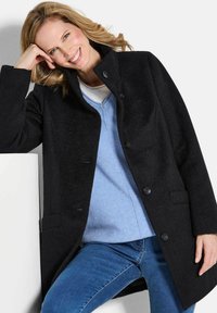 Cappotto di lana nero con colletto alto, bottoni frontali e tasche laterali, indossato sopra un maglione blu e jeans di denim. Texture liscia.