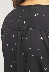 Haut noir en tissu lisse présentant un motif de petits oiseaux blancs. Le modèle inclut un pli au dos pour un détail supplémentaire.