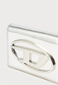 Sidabrinė stačiakampė piniginė su lygiu, blizgiu paviršiumi. Yra ryškus ovalo formos išpjovimas ir tekstūruota kraštinė. Minimalistinio dizaino.