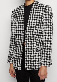 Svart och vit houndstooth-mönstrad kavaj med en strukturerad design, slät textur, öppen front och två framfickor.