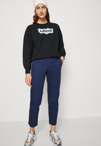 Felpa nera con logo Levi's, pantaloni blu navy e sneakers grigie. Il modello indossa un berretto di maglia bianco. Outfit casual con vestibilità rilassata.