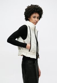 Gesteppte Weste in Creme mit schwarzen wellenförmigen Mustern. Verfügt über einen hohen Kragen, Reißverschluss und gerafften Saum. Wird über einem schwarzen, gerippten Pullover getragen.