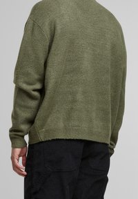 Zelený pletený svetr s uvolněným střihem, kulatým výstřihem a žebrovanými manžetami. Materiál vypadá měkký a strukturovaný, s mírně nerovným lemem.