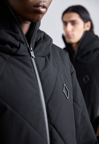 Veste matelassée noire avec un col montant, dotée d'une fermeture éclair à l'avant et d'un petit accent en métal en forme de diamant sur la poitrine.