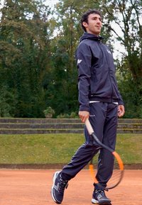 Atleta com uma jaqueta e calças desportivas pretas, segurando uma raquete de ténis com um detalhe laranja, numa quadra de terra batida rodeada de árvores.