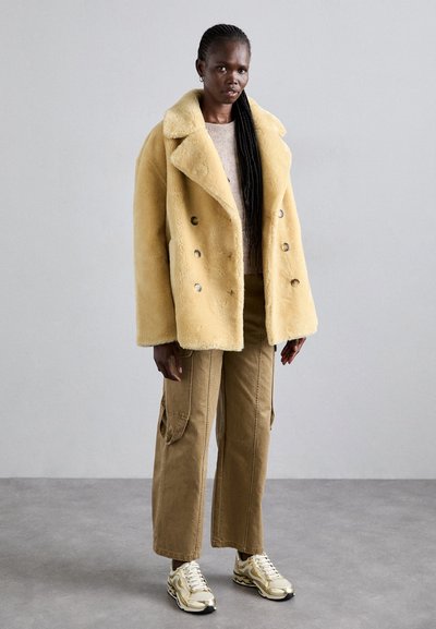 Gelber Kunstpelz-Mantel im Doppelbreasted-Stil mit großem Revers und silbernen Knöpfen, kombiniert mit khakifarbenen Cargo-Hosen und Sneakers mit goldenen Akzenten.