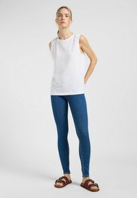 Top blanco sin mangas con escote redondo, combinado con jeans azul ajustados y sandalias marrones con acentos de hardware decorativo.