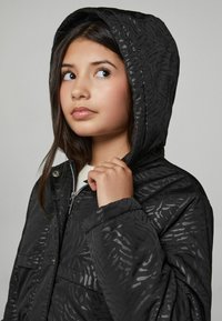 Melna pūkaina jaka ar kapuci, ar diskrētu dzīvnieku rakstu tekstūru, quilted dizainu, rāvējslēdzēju un priekšējo kabatu.