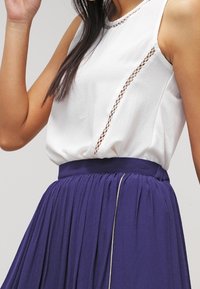 Blusa sin mangas blanca con detalles de encaje, combinada con una falda morada fluida que presenta un acento dorado a lo largo de la cinturilla.