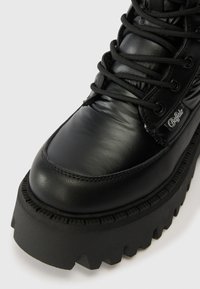 Črne masivne platform škornje z gladkim usnjem, okroglo noso, izrazitimi čipkami in teksturiranim gumijastim podplatom z izstopajočimi robovi.