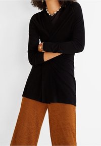 Personne portant un haut noir à manches longues avec un devant croisé et un pantalon large couleur rouille texturé, accessoirisé d’un collier et de bracelets coquillages.
